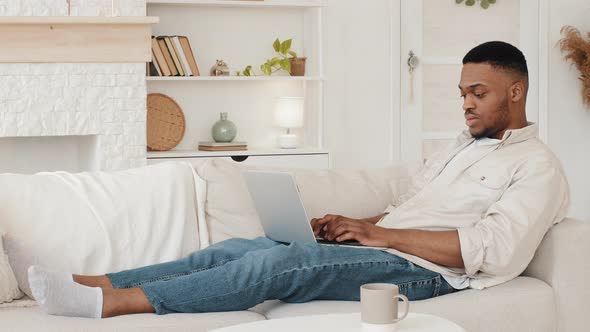 Focused African Guy Student Lying on Couch with Laptop Typing Browsing on Web Study Remotely From alt