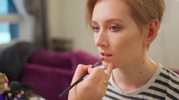 a Young Woman with a Short Haircut Sits and a Makeup Artist Applies Lipstick to Her Lips with a alt