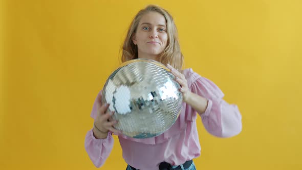 Slow Motion Portrait of Young Woman Holding Disco Ball Dancing on Yellow Background alt