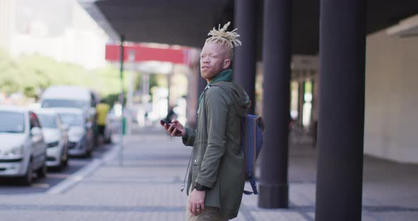 Thoughtful albino african american man with dreadlocks walking on street using smartphone alt