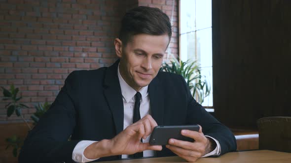 Portrait of Smiling Businessman Man Watching Funny Videos on Smartphone While Sitting in Modern