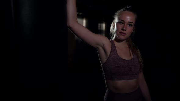 Confident Athletic Woman Standing at Punching Bag in Darkness Looking at Camera alt