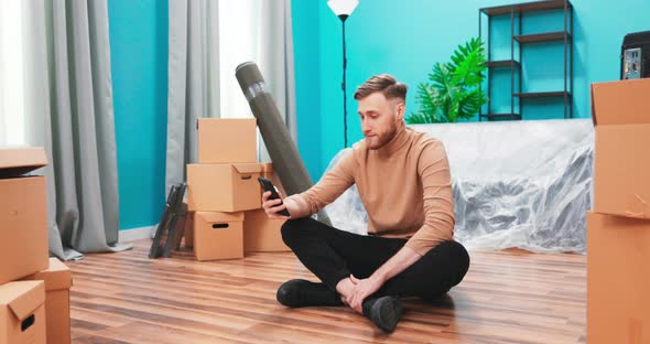 Young Man are Using Phone Amid Boxes in a New House alt