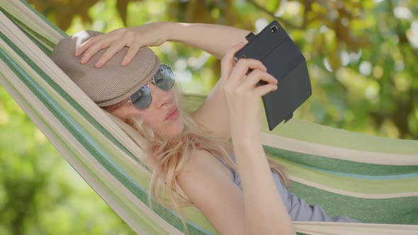 Smiling blonde woman with sunglasses using smartphone, lying relaxing on the hammock in the garden alt