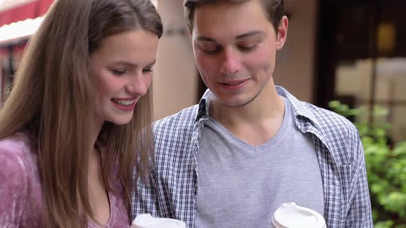 Happy Couple Using Mobile Phone And Drinking Coffee Outdoors