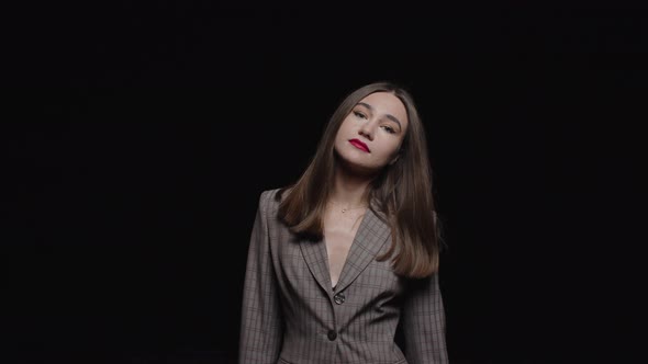 A beautiful girl in a jacket looks into the camera and sways to the sides on a black background.