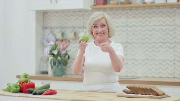 Healthy Senior Old Woman Showing Thumbs Up While Holding Apple in Kitchen alt