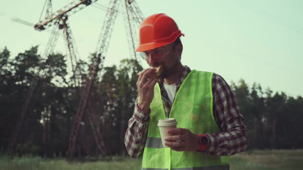 Hungry Worker in Overalls in Electric Power Business Eats Sandwich and Drinks Takeaway Coffee at alt