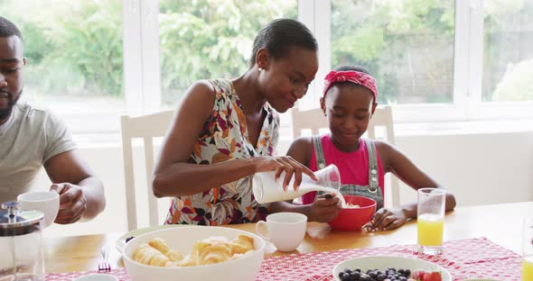 African american mother pouring milk in daughter's cereal bowl at home alt