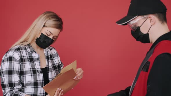 Woman in a Mask Is Receiving a Parcel of Craft Paper Envelopes From a Deliveryman alt