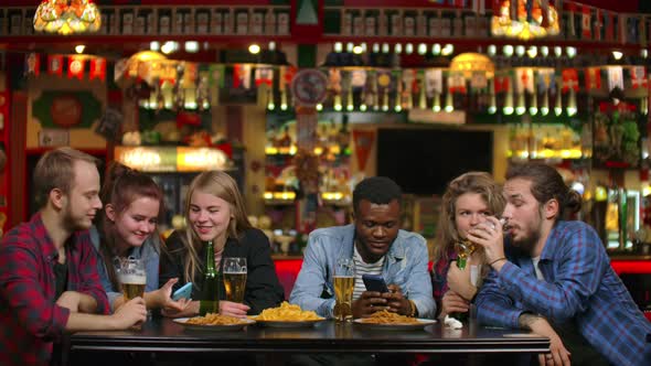 Cheerful Students Sitting at a Table in a Bar Drinking Beer, Eating Chips and Watching Photos on a alt