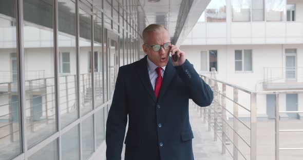 Mature Angry Businessman Talking on Mobile Phone Walking Outside Business Center in City alt