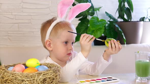 A little boy in rabbit ears enthusiastically paints Easter eggs with paint.