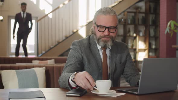 Senior Businessman Drinking Coffee and Working on Laptop in Restaurant alt