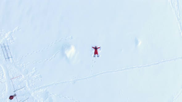 Aerial Camera Rises Up, a Top View of a Girl in a Red Fluff Lying and Making a Snow Angel alt