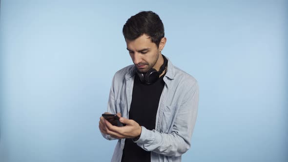 Young Confident Man Holding Phone and Scroll Screen or Typing Message Isolated on Blue Background alt