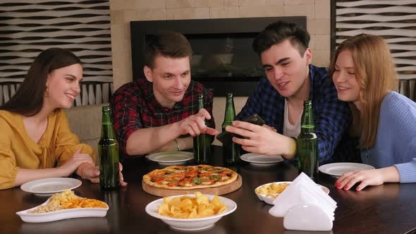 Friends Relax in a Pizzeria with Beer, They Look at the Phone Together and Laugh alt