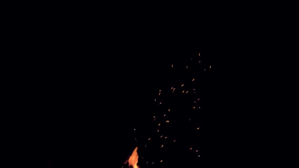 Glowing embers drift up into the darkness of the night sky from a campfire. alt
