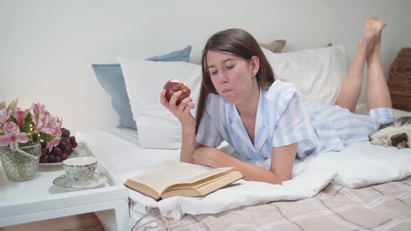 A Woman Eats an Apple and Reads a Book While Lying in Bed