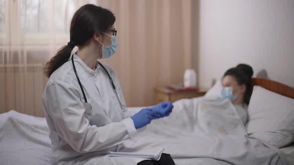 Concentrated Female Doctor Putting on Gloves Looking Back at Blurred Ill Woman Lying in Bed in alt