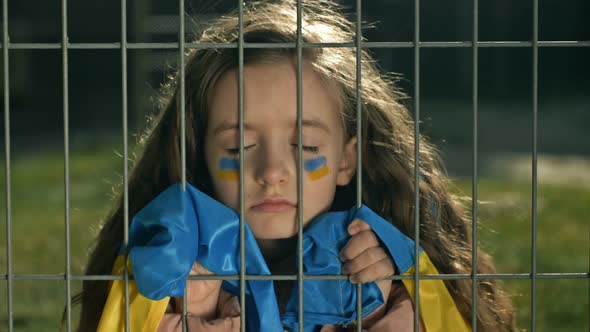 Little Girl is Covered with a Ukrainian Flag with Her Eyes Closed Behind a Metal Grill