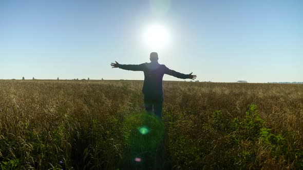 Farmer Businessman with Raised Hands Walks Through the Wheat Field at Sunrise alt