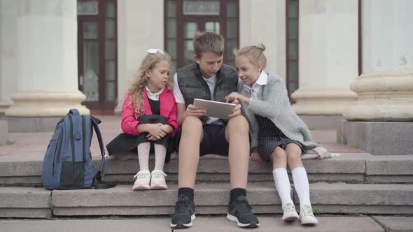Wide Shot of Three Classmates Sitting on Break Outdoors and Surfing Internet on Tablet. Caucasian alt
