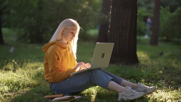 A Young Woman in Jeans and a Bright Yellow Sweatshirt with a Laptop Sits on the Green Grass alt