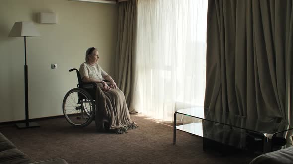 Senior Woman Sit on Wheelchair and Looks Through the Window alt