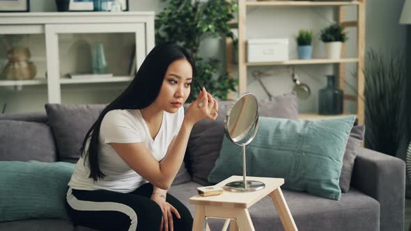 Beautiful Asian Girl Is Putting on Make Up Using Mascara and Face Powder Looking at Mirror Sitting alt
