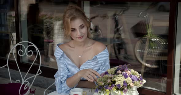 Young Girl Sitting In A Cafe And Admires A Bouquet Of Flowers alt