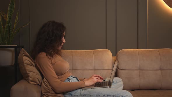 Young Woman Sitting on Couch Using Laptop Looking at Screen Typing Message Lady Chatting on Computer alt