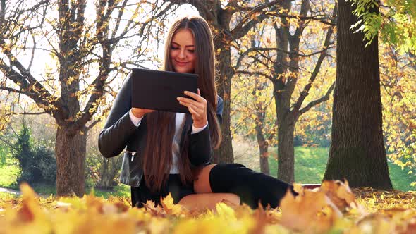 Young Beautiful Woman Lies on the Foliage in Autumn Park and Works on Tablet alt
