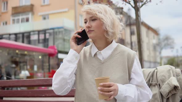 Young Blonde Business Woman in a Beige Vest and White Blouse Sits on a Bench in the Middle of the alt