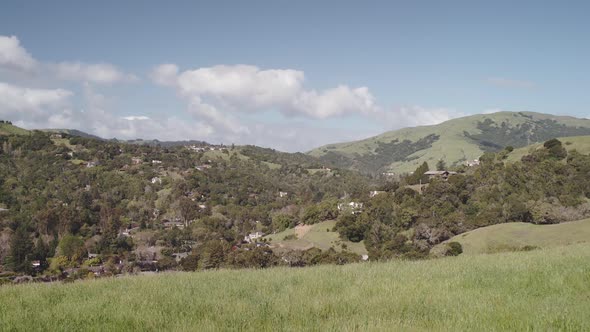 The camera cranes down over an idyllic green valley. Houses and clouds can be seen in the distance. alt