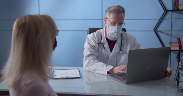 Professional Serious Focused Middle Aged Hoary Physician in Medical Mask and Coat Sitting at Table