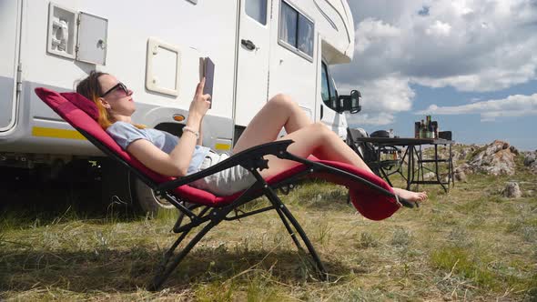 Woman Sitting On Folding Chair Reading Book On Camp Site Next To Camper Rv