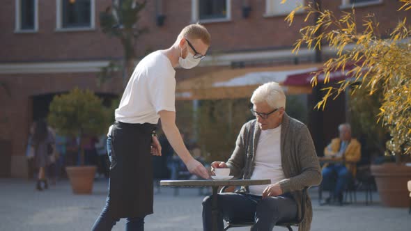 Waiter Wearing Face Mask Serving Coffee for Senior Man in Street Coffee Shop alt