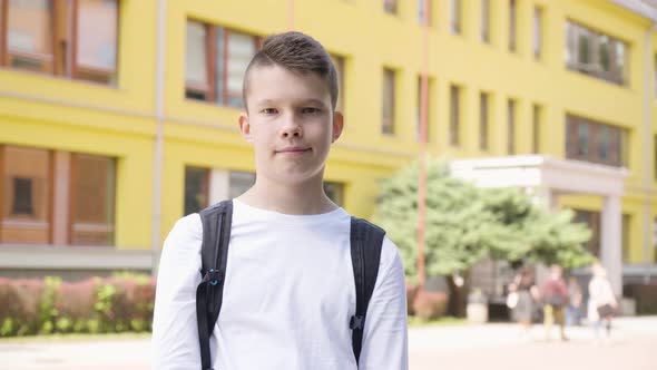 A Caucasian Teenage Boy Shows a Thumb Up to the Camera with a Smile  a School in the Background