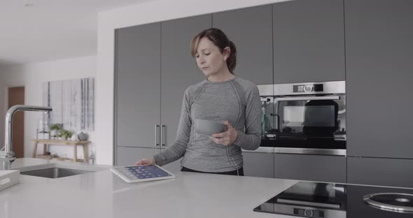 Active Mature woman eating healthy breakfast in kitchen and checking email on digital tablet