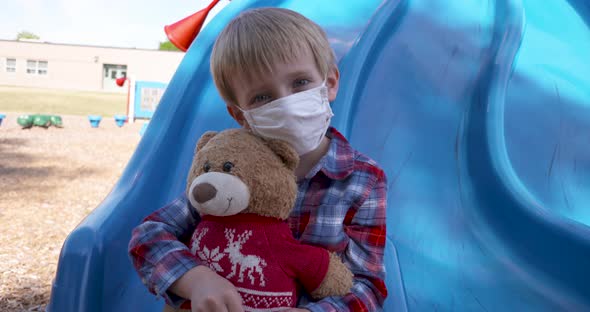 Little boy in a mask cuddling his teddy bear at a playground. alt
