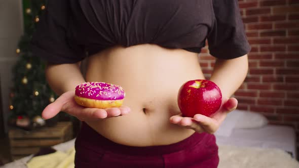 Hands of Plump Woman Holding an Appetizing Donut and Fresh Apple Against Background of Large Belly alt