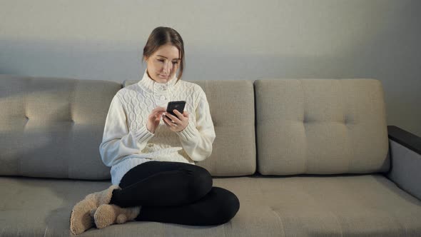 Pretty Girl in Sweater Sitting on Sofa and Read Messages on the Phone alt