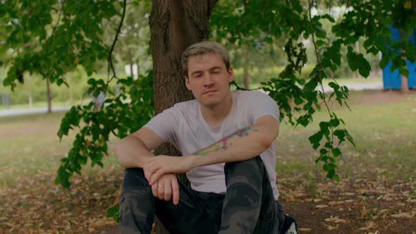 Close Up of Young Man Rests Sitting Near Tree in Parkland alt