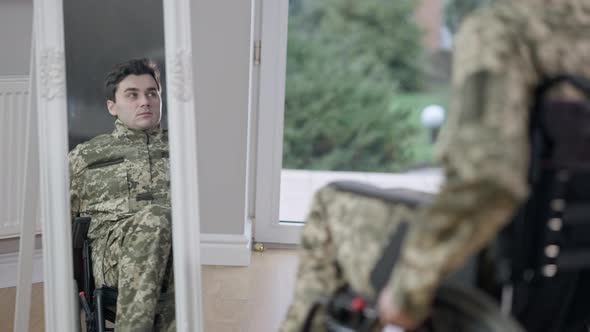 Sad Young Paralyzed Man in Camouflage Riding on Wheelchair From Mirror to Glass Door alt