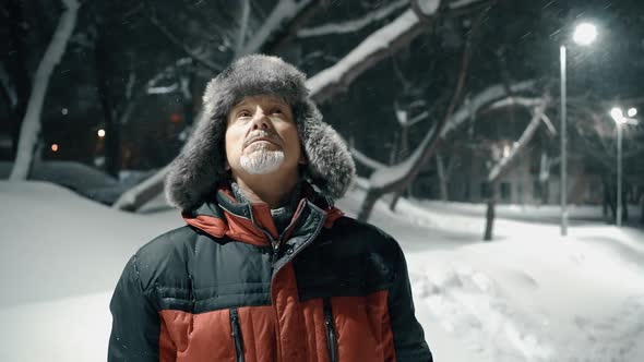 An Elderly Man with a Gray Beard in a Winter Hat is Walking Late in the Evening on a Snowcovered