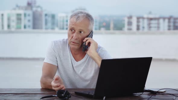 An Elderly Whitehaired Male Businessman is Talking on the Phone Sitting at a Laptop in the Evening