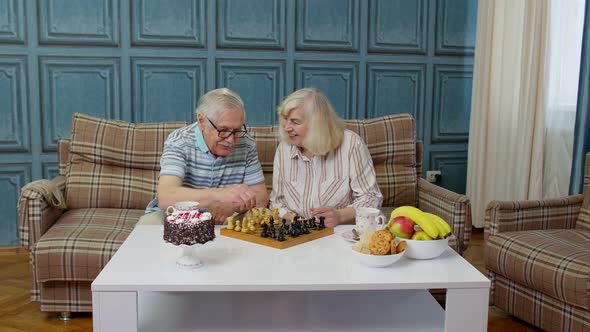 Retired Senior Couple Talking Drinking Tea Playing Chess in Modern Living Home Room Lounge Together alt