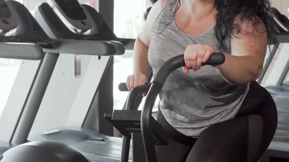 Lovely Cheerful Plus Size Woman Smiling To the Camera Exercising on Air Bike alt