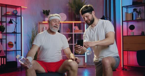 Young and Old Men Doing Break in Joint Home Sports Training and Watching Funny Videos on Mobile alt